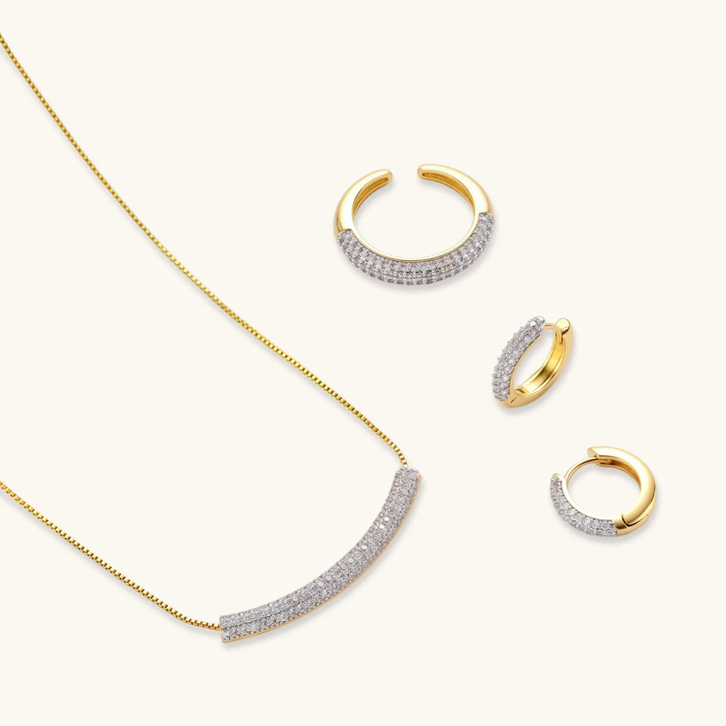 Gold-plated curved necklace, hoop earrings, and adjustable ring with pavé cubic zirconia stones on cream background.
