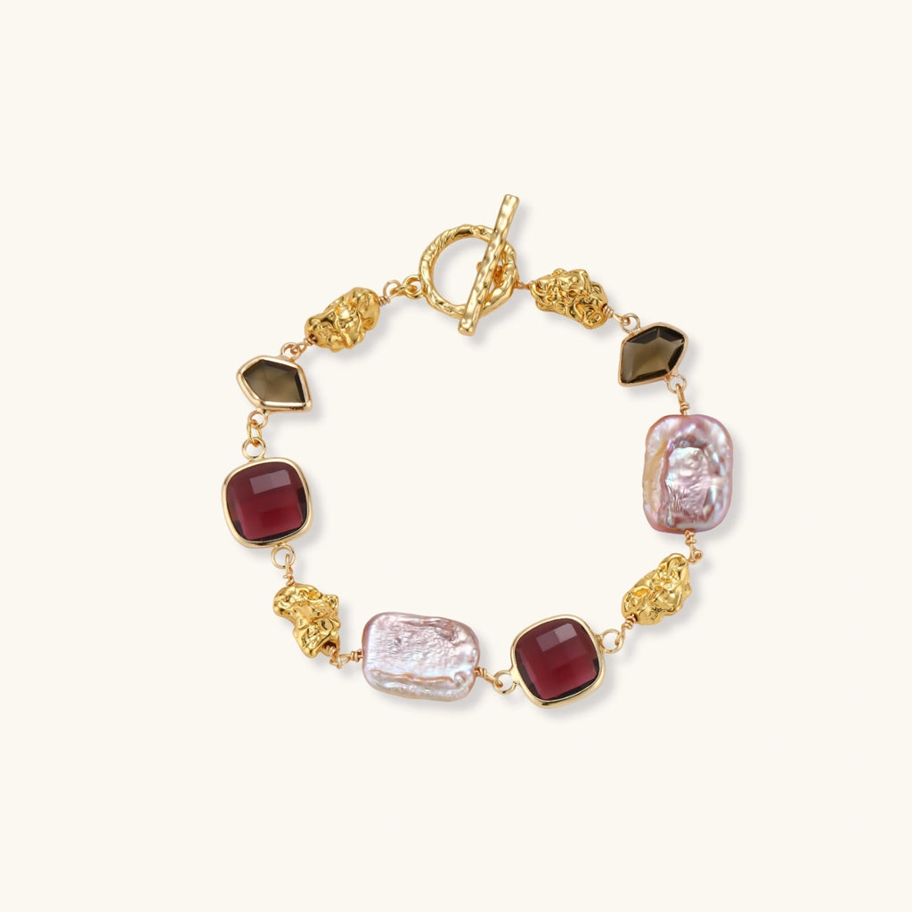 18k gold-plated copper bracelet with burgundy stones, smoky quartz, baroque pearls and ornate golden charms on white background.