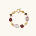 18k gold-plated copper bracelet with burgundy stones, smoky quartz, baroque pearls and ornate golden charms on white background.