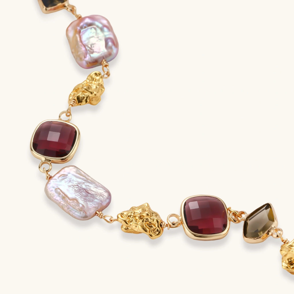 18k gold-plated copper bracelet with burgundy stones, smoky quartz, baroque pearls and ornate golden charms on white background.