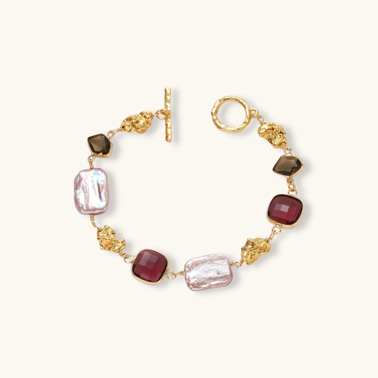 18k gold-plated copper bracelet with burgundy stones, smoky quartz, baroque pearls and ornate golden charms on white background.